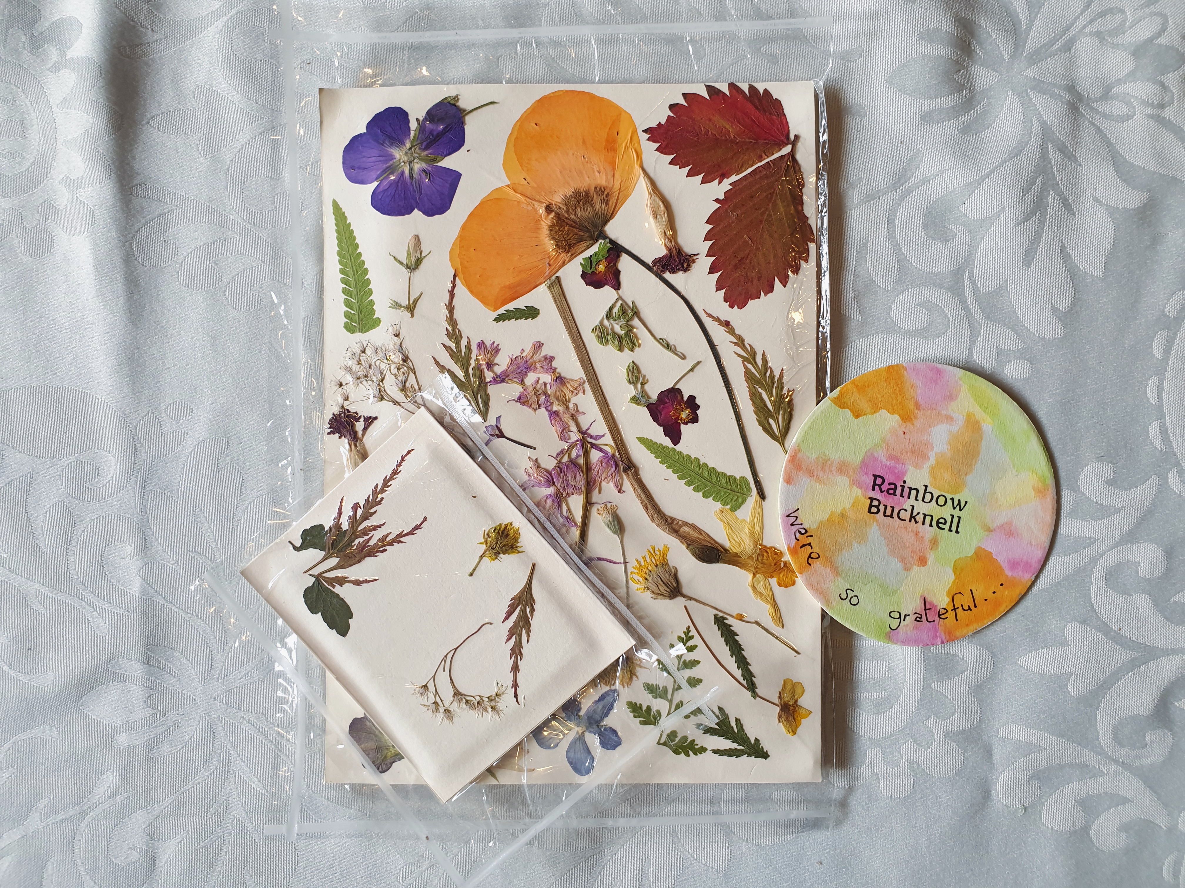 Photo containing a pack of pressed flowers/foliage, a small miniature version, and a brightly coloured round piece of card with ‘Rainbow Bucknell’ stamped on it along with handwritten wording of “We’re so grateful…”